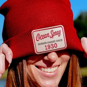 Smiling person pulling a red knit beanie with an Ocean Spray "Farmer-Owned Since 1930" patch down over their eyes